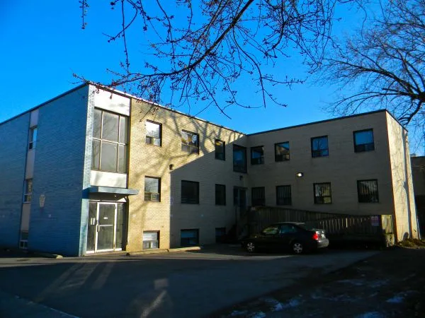 a photograph of a two-story brick building with a blue sky and bare tree branches overhead, a dark car parked in front, and…