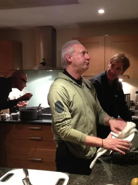 A man in a green hoodie stands in a kitchen, looking upward with a slightly annoyed expression while drying dishes. Behind…
