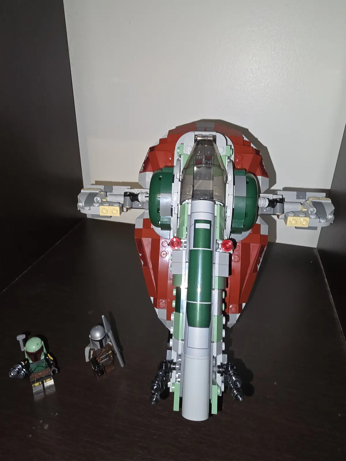 A large, red, green, and gray LEGO model of a Star Wars starfighter sits on a dark wooden surface, with two LEGO minifigures…