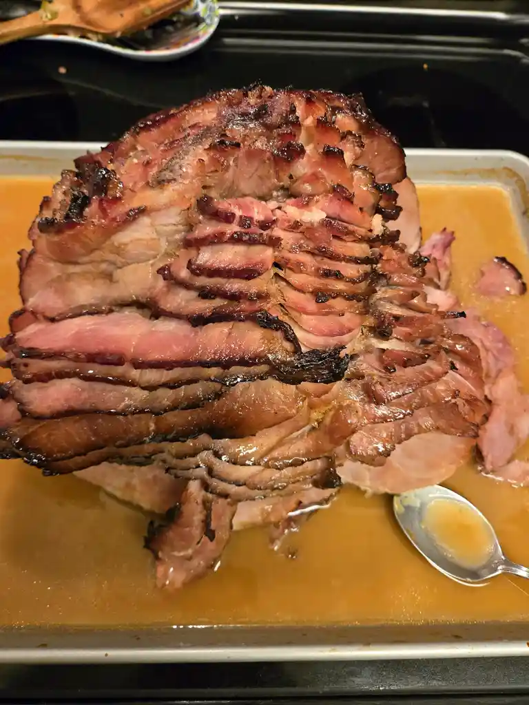A close-up photo shows a large, partially sliced roasted ham on a baking tray, surrounded by its own brownish cooking…