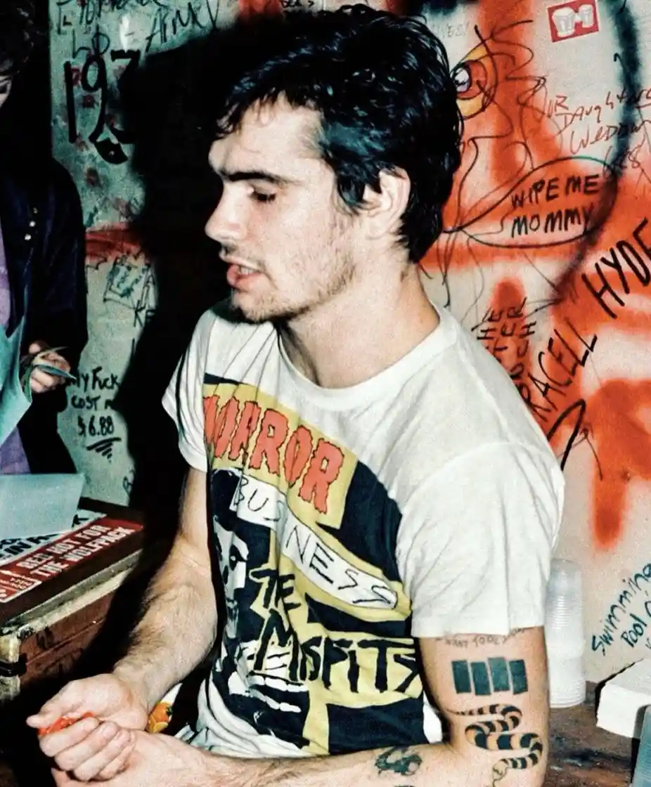A man with dark hair and tattoos on his arms sits in front of a graffiti-covered wall. He wears a graphic t-shirt with…