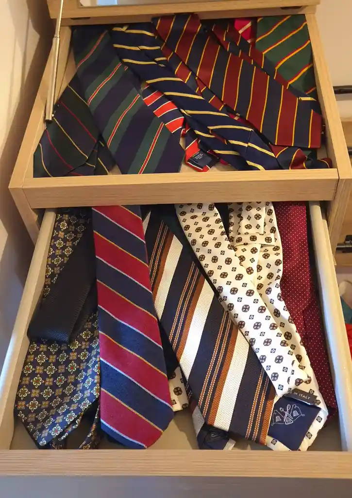 A wooden drawer is open, revealing two compartments filled with numerous colorful neckties. The top drawer is overflowing…