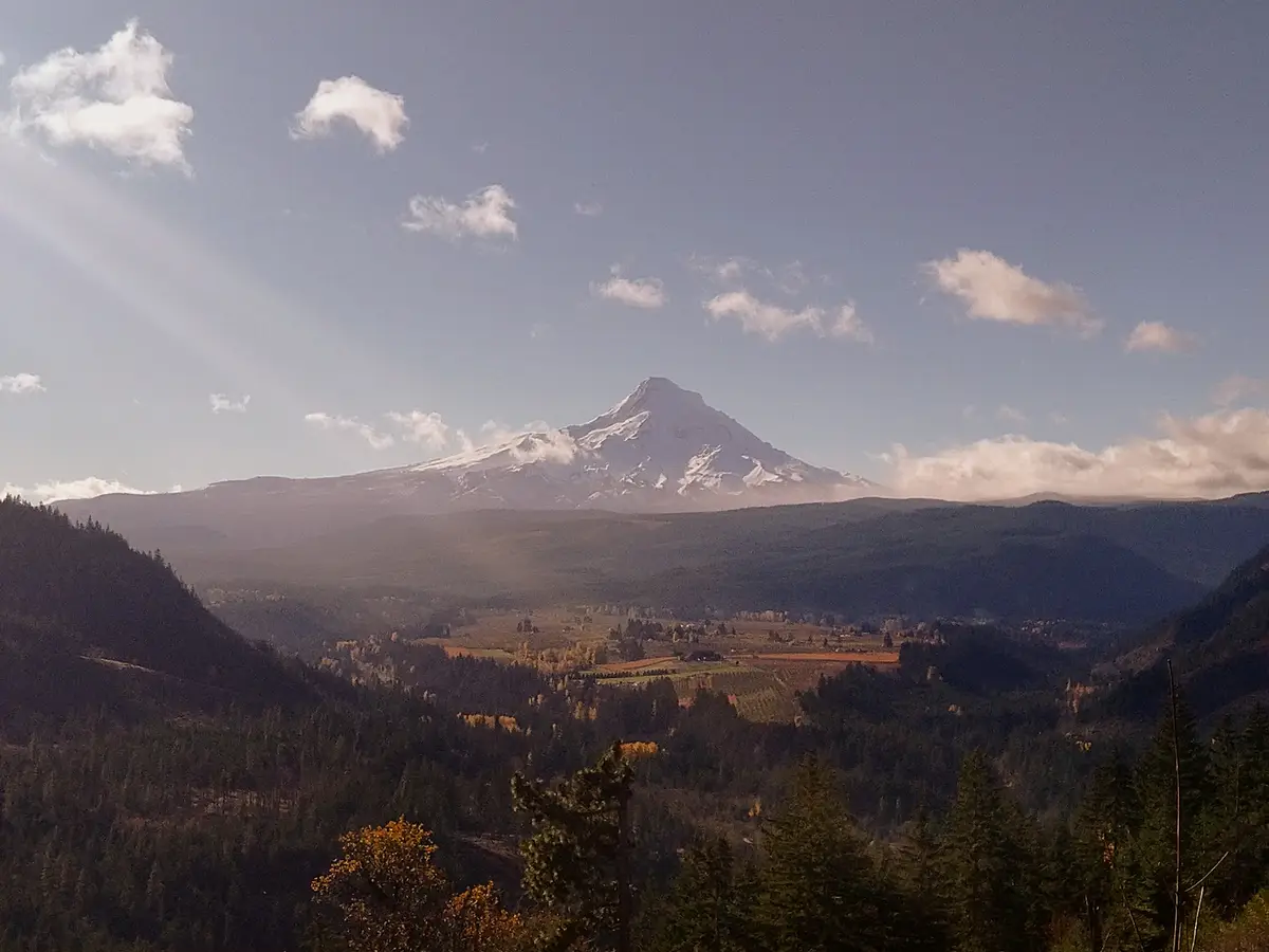 A scenic landscape photo shows a snow-capped mountain, likely Mount Hood, rising above a valley with forests and fields…