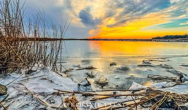 A scenic winter landscape shows a frozen lake at sunset, with ice chunks, snow, and reeds in the foreground. The sky is…