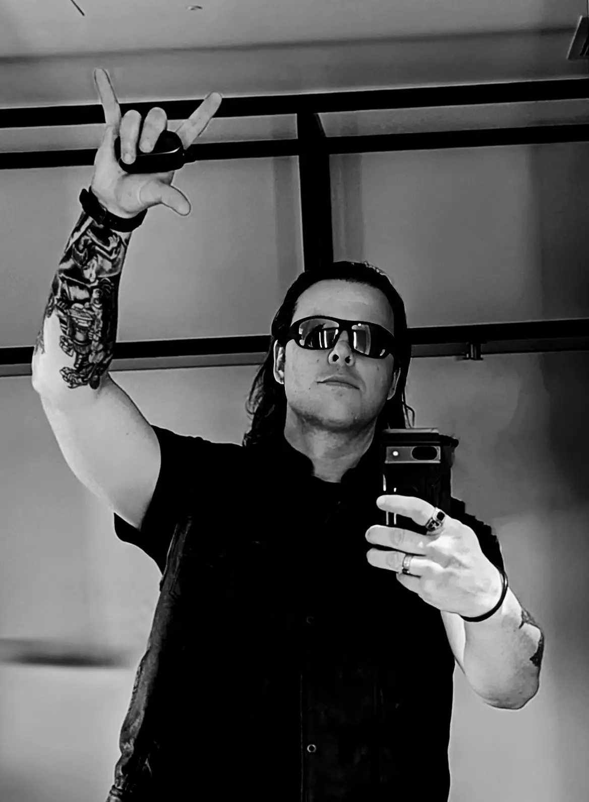 A black-and-white mirror selfie of a man with long hair and sunglasses, holding a phone in one hand and making a rock hand…