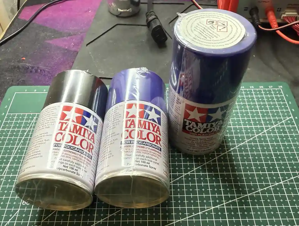Three Tamiya brand spray paint cans, labeled 'FOR POLYCARBONATE' and 'FOR PLASTICS', are shown lying on their sides on a…