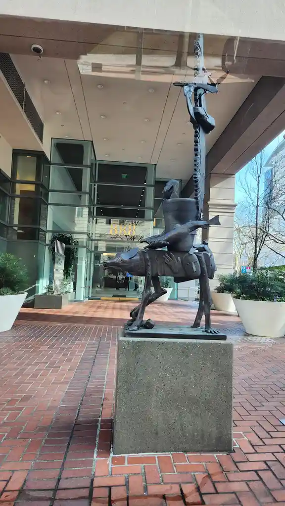 A bronze sculpture of a stylized dog carrying a bucket, fish, and other figures stands on a concrete pedestal in front of a…