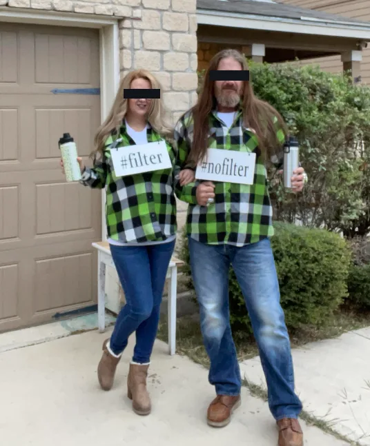 A man and woman stand outside a house wearing matching green and black plaid shirts, holding signs that read '#filter' and…