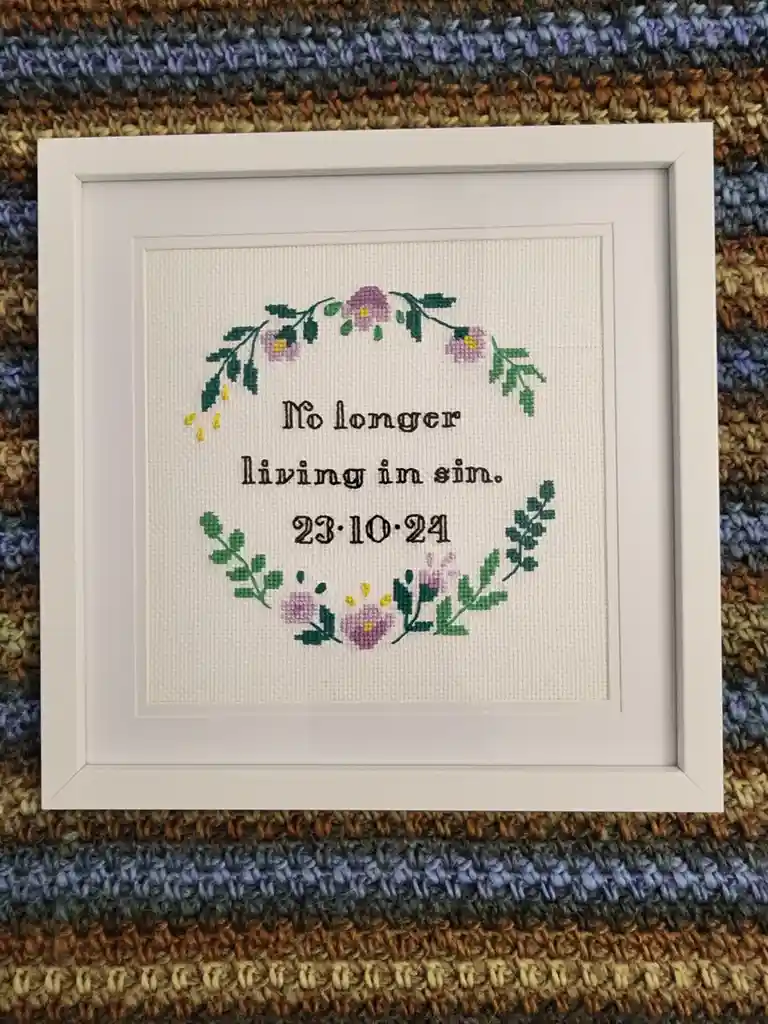 A white-framed cross-stitch piece on white fabric displays the text 'No longer living in sin. 23.10.24' in black thread…