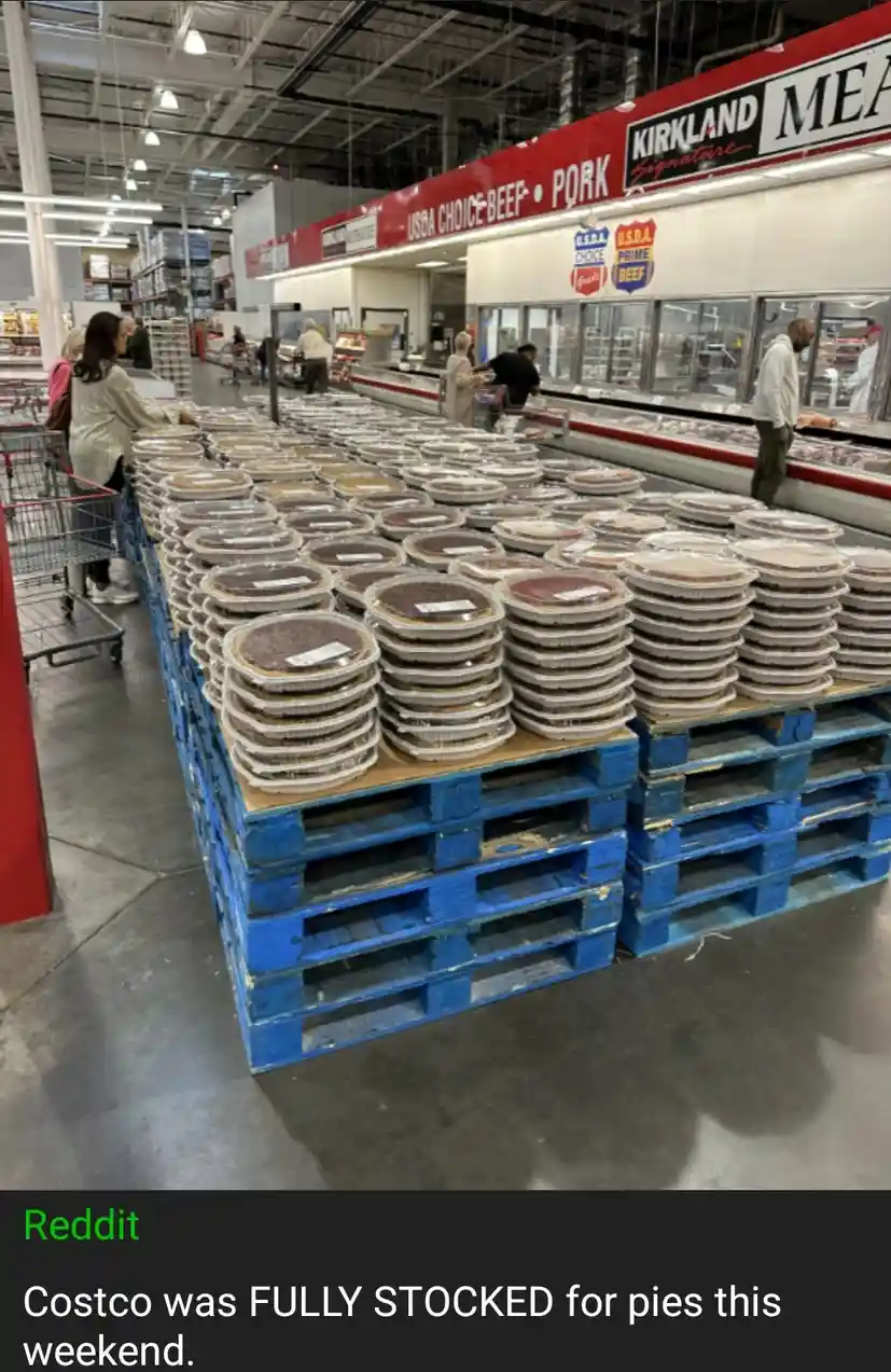 A Costco meat aisle is visually replaced by massive stacks of pre-packaged pies on blue pallets, with shoppers browsing…