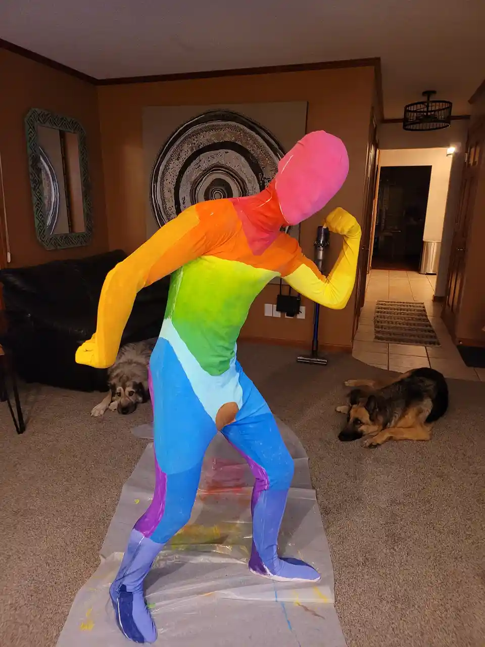 A person wearing a full-body, brightly colored rainbow suit stands in a living room, flexing their bicep while standing on a…