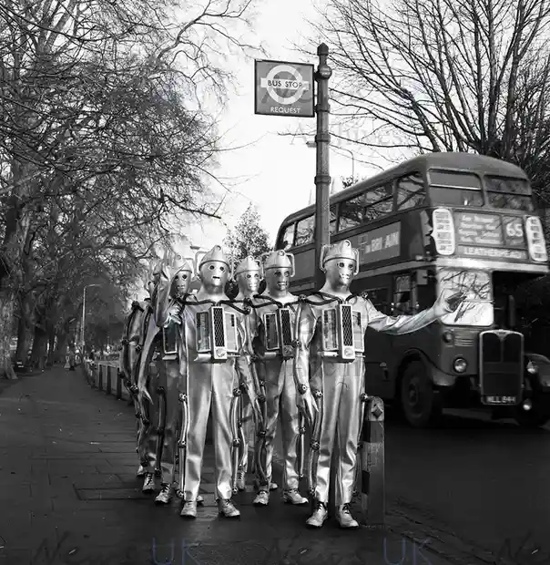 A black-and-white photo shows a line of people in silver, metallic, robot-like costumes standing at a bus stop. One person…