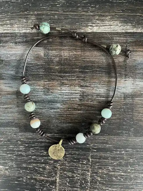 A brown leather cord bracelet with various green and white beads and a small gold coin charm lies on a dark, weathered…