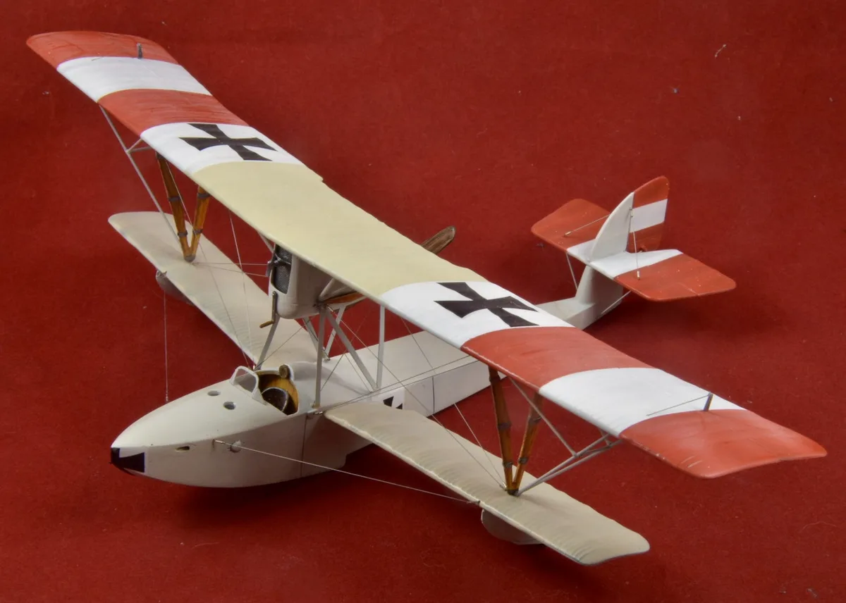 A detailed model of a biplane with red and white striped wings and black crosses, displayed against a solid red background…