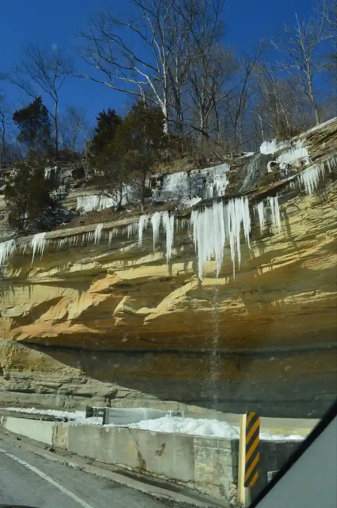 A photo taken from inside a car shows a steep, layered cliff face with numerous long icicles hanging from its top edge. The…