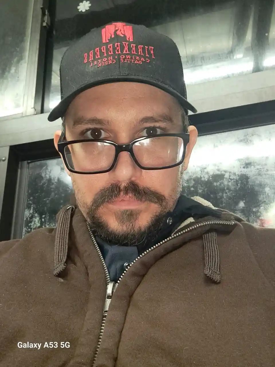 A man wearing a black baseball cap with red 'FIREKEEPERS CASINO HOTEL BATTLE CREEK' text, black-rimmed glasses, and a brown…