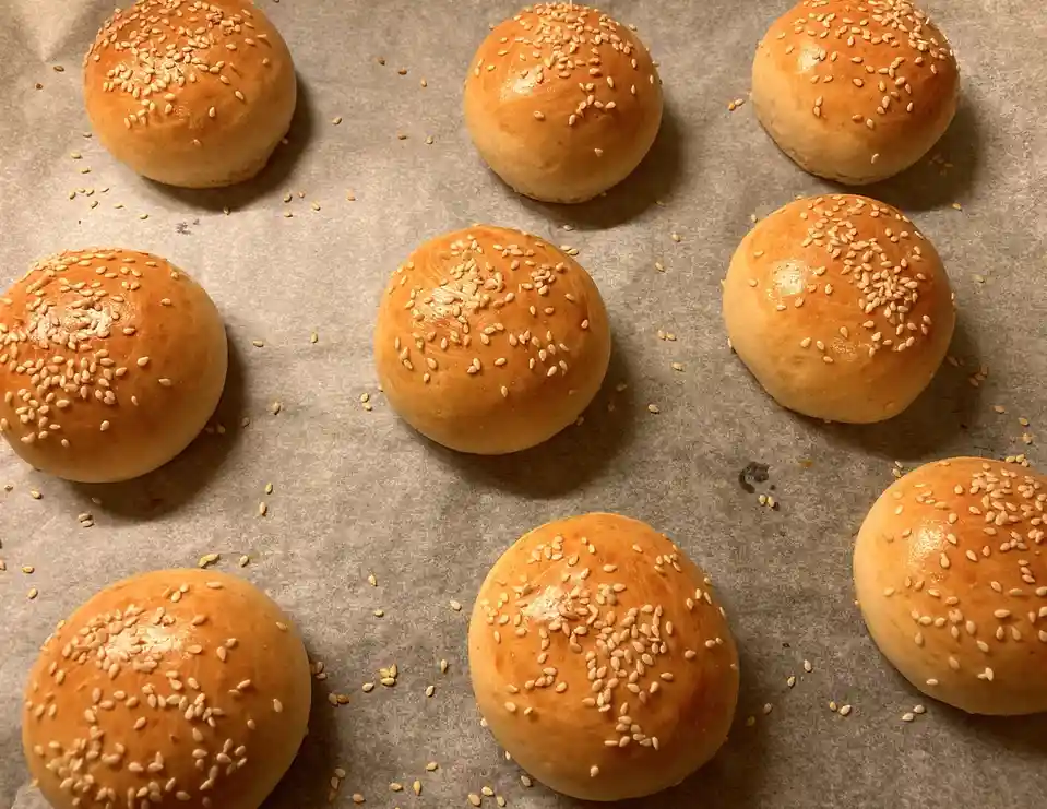 A close-up photo shows nine freshly baked, golden-brown sesame seed buns arranged on parchment paper. The buns are round…