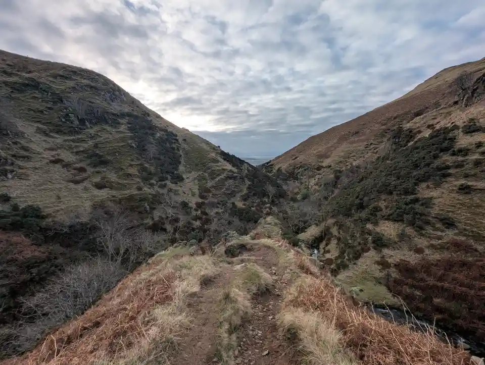 A narrow dirt trail winds down a steep, grassy mountain valley under a cloudy sky, flanked by rugged hillsides with sparse…