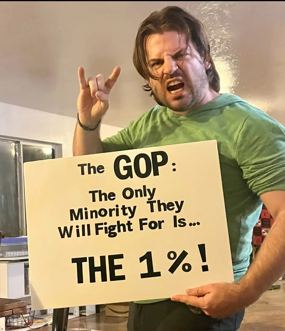 A man with long brown hair and a green shirt is holding a white sign with black text that reads 'The GOP: The Only Minority…