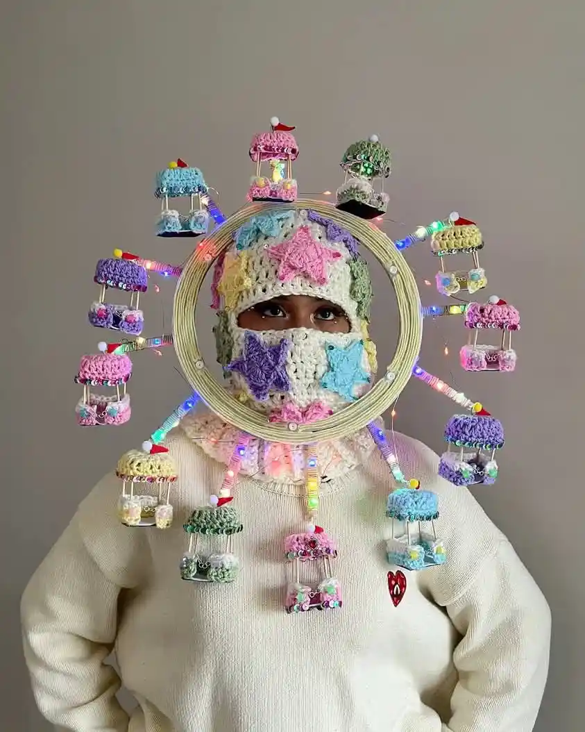 A person wearing a cream-colored sweater and a crocheted balaclava with colorful star appliqués stands against a plain…