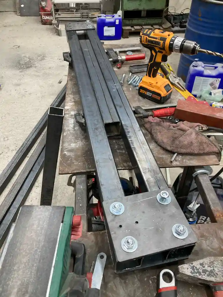 A workshop scene shows a makeshift drill press constructed from metal square tubing, bolts, and a yellow and black cordless…