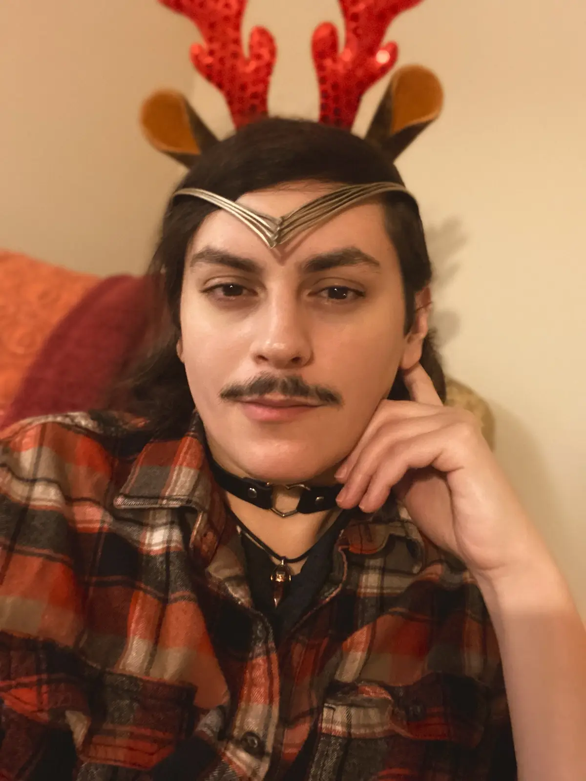A person with long dark hair and a mustache wears a red sequined reindeer antler headband, a silver Elven-style brow band…