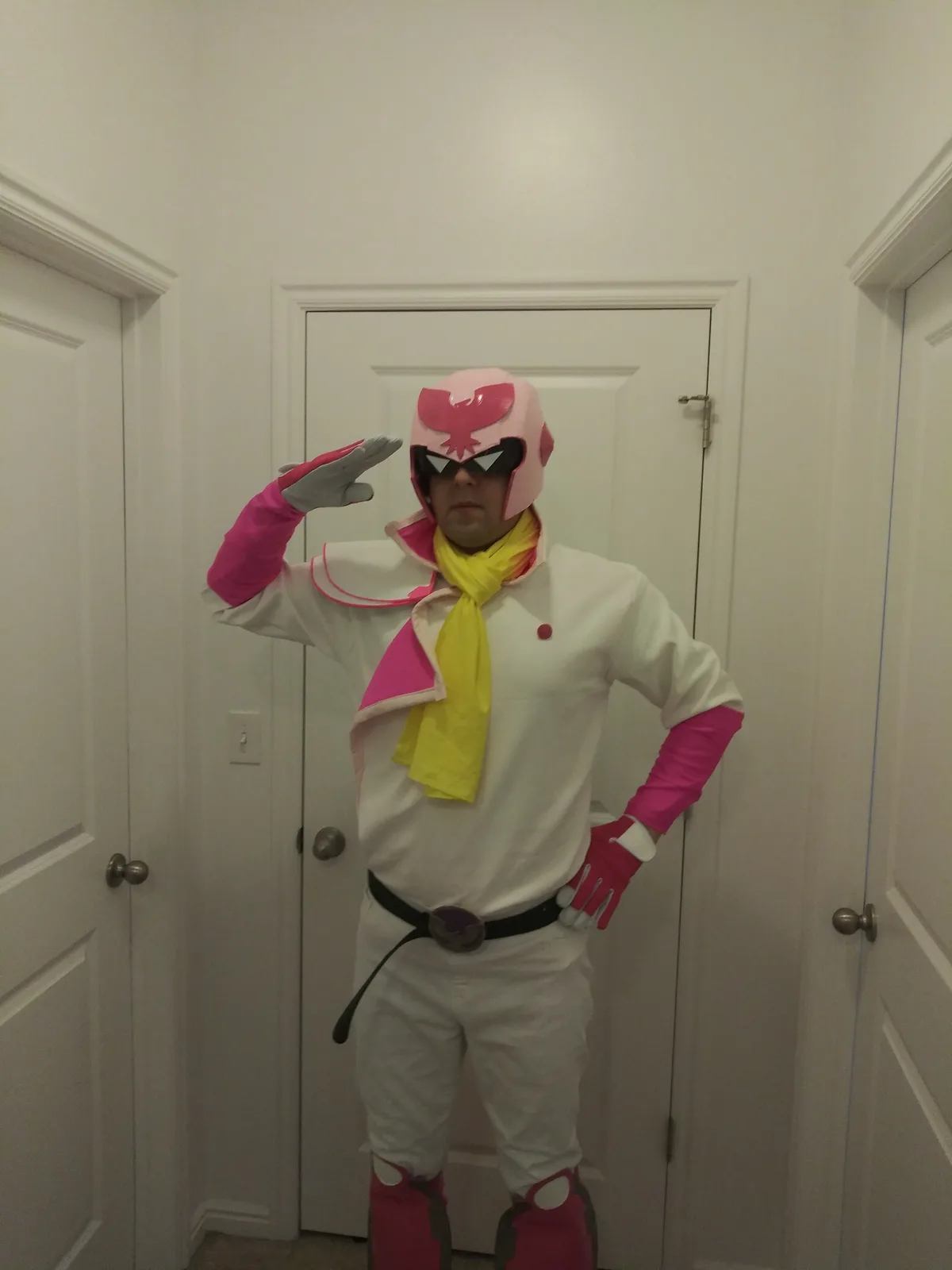 A person in a full-body pink and white superhero costume with a bird emblem on the helmet, yellow scarf, and black belt…