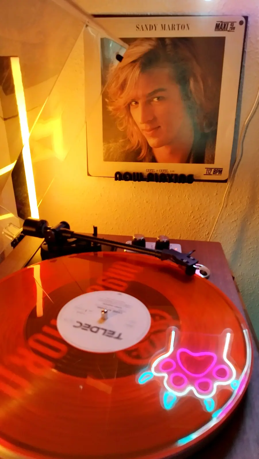 A close-up shot of a red vinyl record spinning on a turntable, with a glowing pink and blue neon paw print sticker on its…