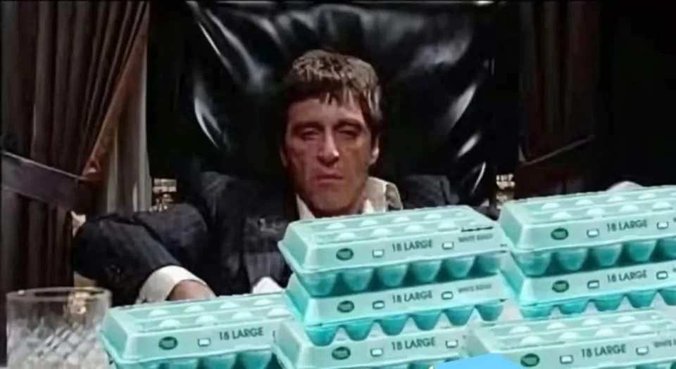 A man with a stern expression sits in a black leather chair behind a table piled with multiple stacks of light blue egg…