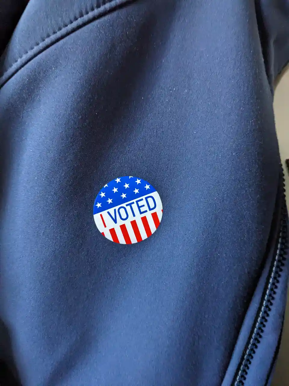 A close-up photo shows a circular 'I VOTED' sticker with American flag design pinned to a dark blue jacket. The sticker…