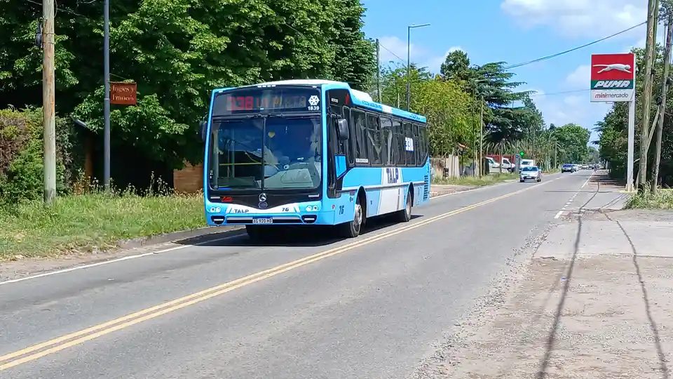 A blue and white public bus, route 338 to La Plata, drives down a sunny road with trees and a PUMA minimercado sign visible…