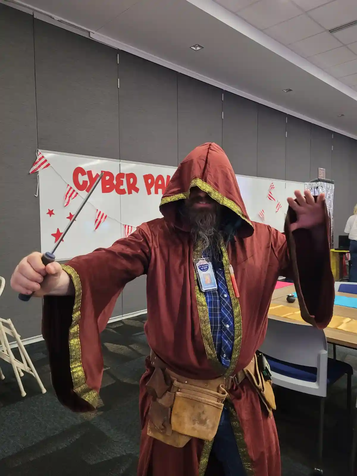 A man in a red hooded robe with gold trim, holding a screwdriver like a wand, poses dramatically in an office setting with a…