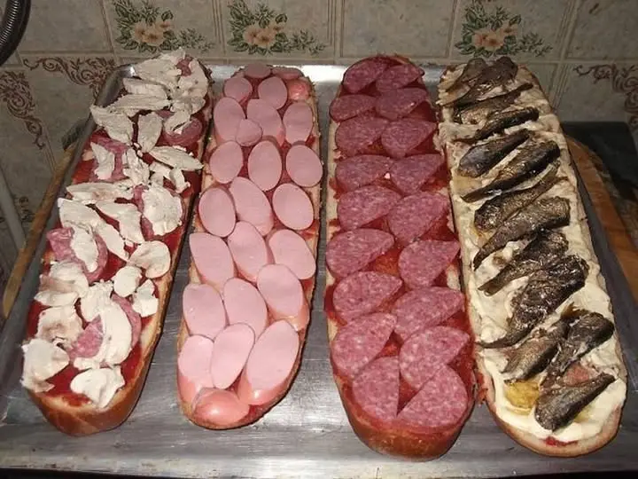 Four long submarine sandwiches lined up on a metal tray, each with a different topping: one with chopped white meat and…