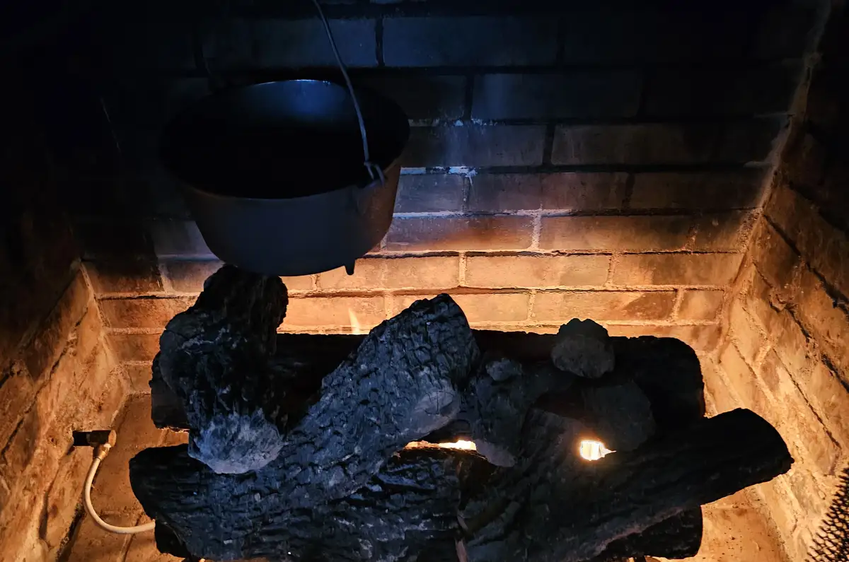 A black cast iron pot hangs over a fireplace with glowing embers and charred logs, set against a brick wall. The overlaid…
