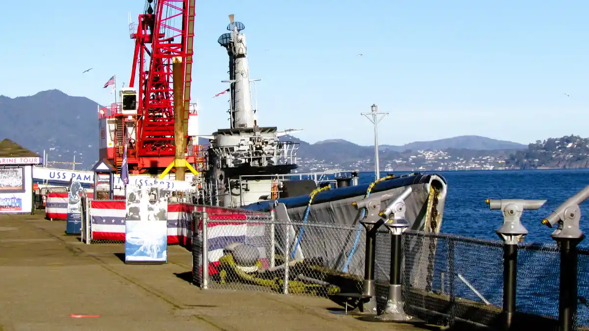 A photo of the USS Pampanito submarine museum docked at a pier, with a red crane structure nearby, American flags, and…