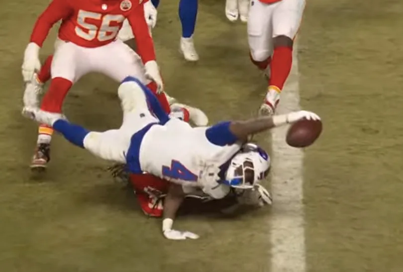 A football player in a white and blue uniform is falling backward on a field, arms outstretched, with the ball flying from…