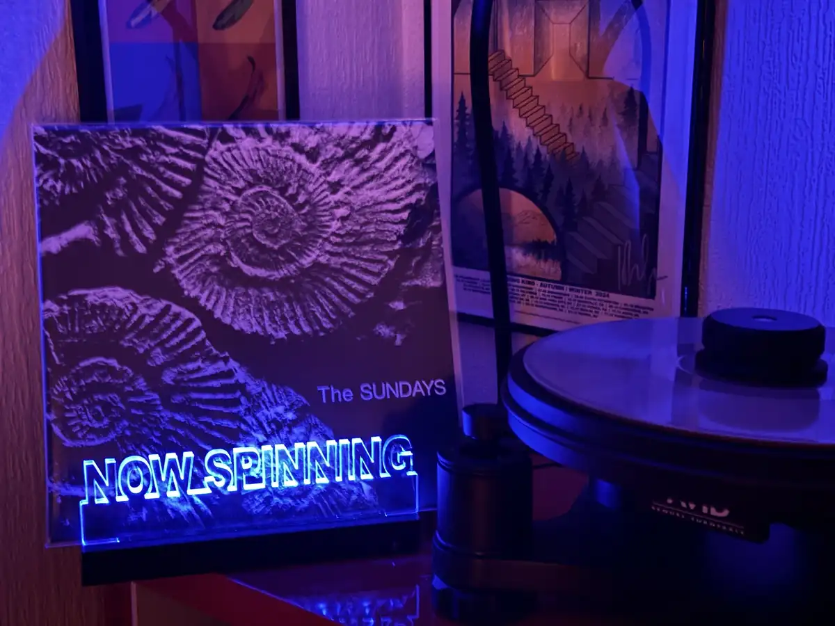 A vinyl record player sits next to a glowing 'NOW SPINNING' sign that displays 'The SUNDAYS' album art featuring ammonite…