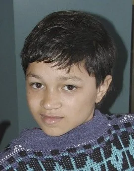 A young person with short dark hair and a purple and teal patterned sweater looks directly at the camera with a neutral…
