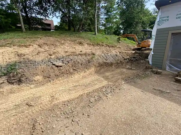 An outdoor construction site shows a partially built retaining wall made of gray interlocking stones, with a large, deep…