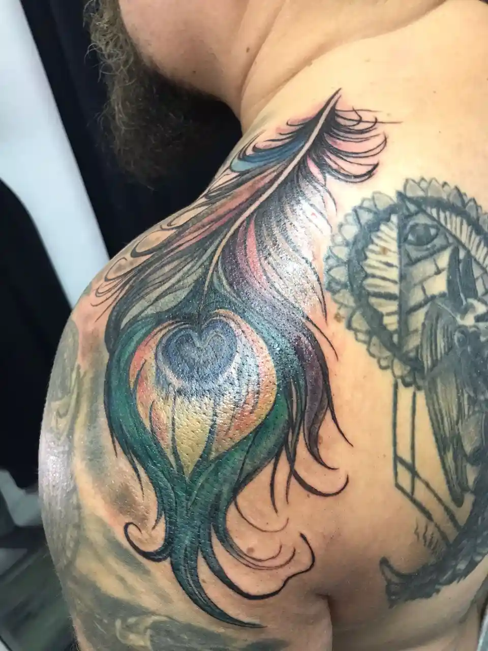 A close-up photo shows a person's shoulder with a large, colorful tattoo of a peacock feather, featuring a heart-shaped eye…