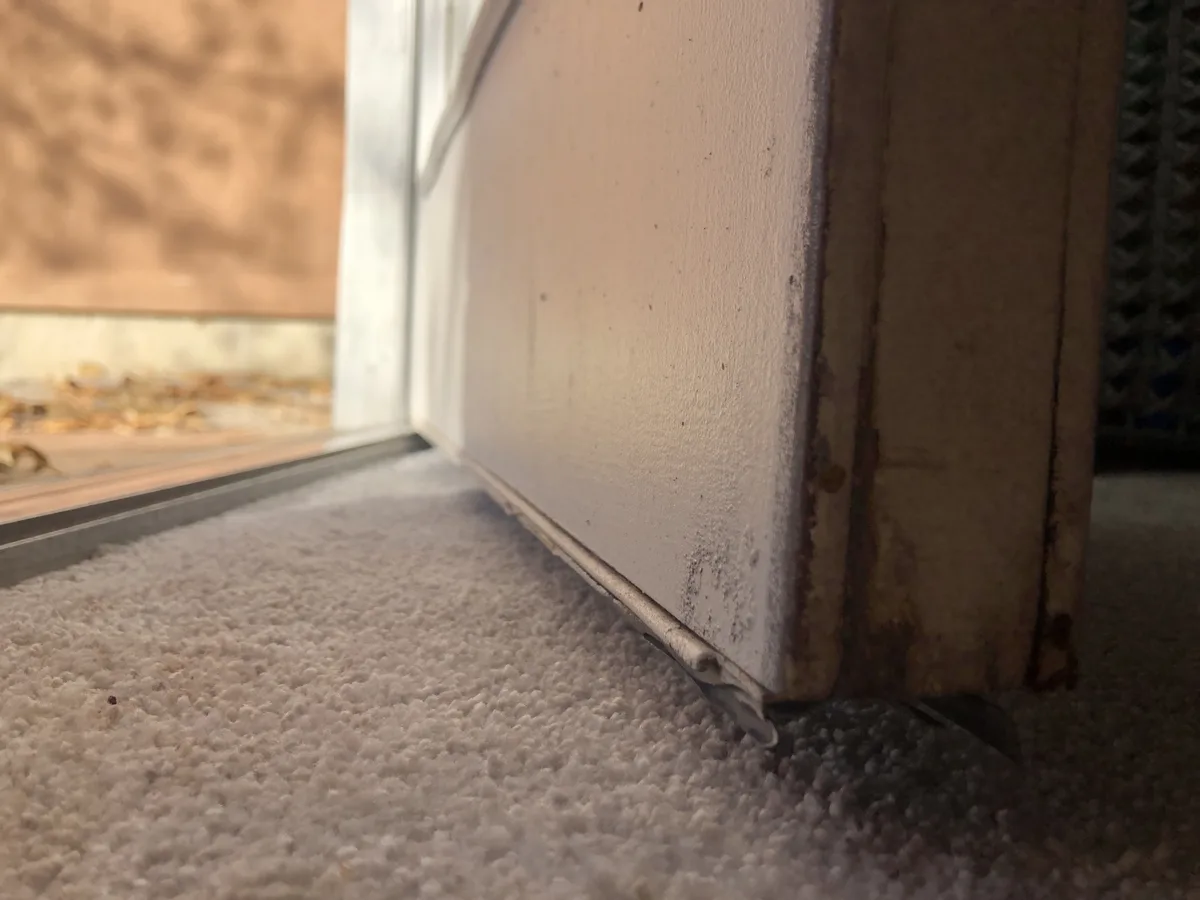A close-up photo shows the bottom of a weathered, rusty door frame resting on a light-colored carpet, with a blurry outdoor…