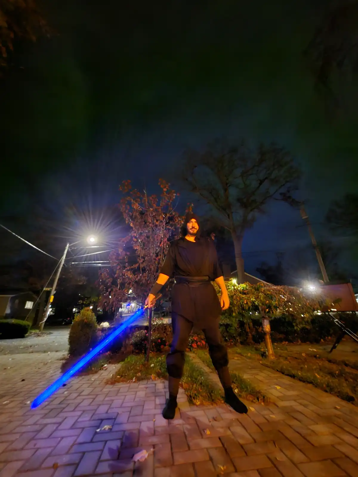 A man in a dark, hooded robe stands on a brick sidewalk at night, holding a glowing blue lightsaber. Streetlights and trees…