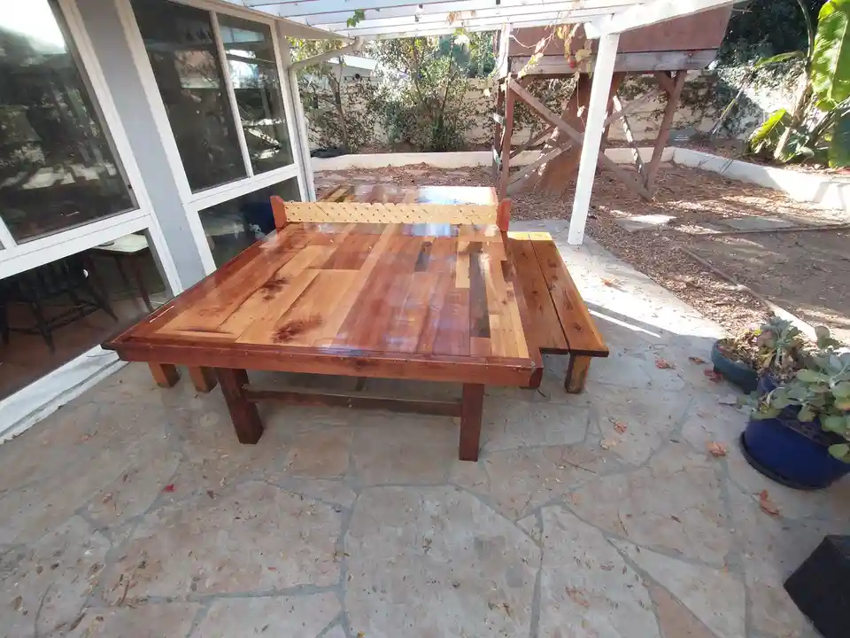 A custom-built wooden ping pong table with attached benches sits on a stone patio under a pergola, with a glass door and…