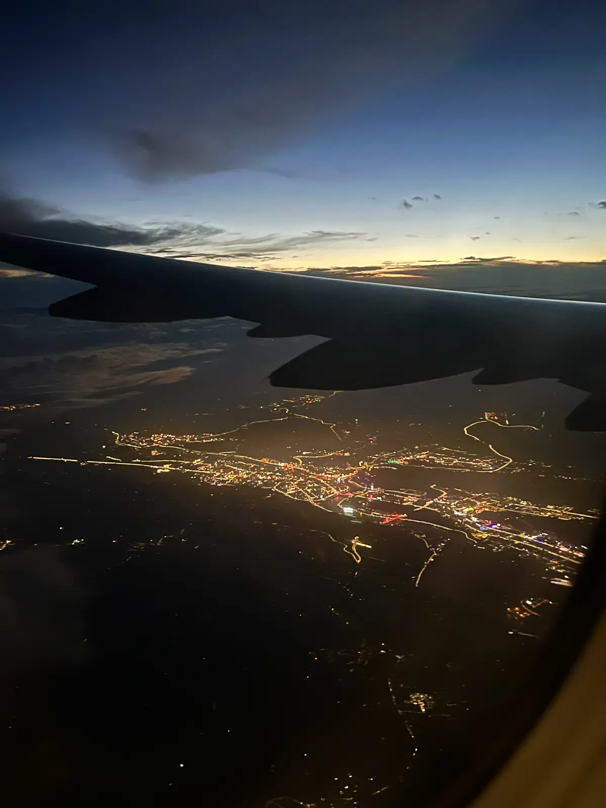 An aerial view from an airplane window at dusk or dawn, showing the silhouette of the aircraft's wing against a gradient sky…