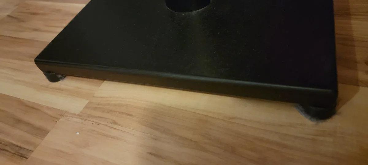 A close-up photo shows the bottom corner of a black electronic device resting on a light-colored wooden floor, with visible…