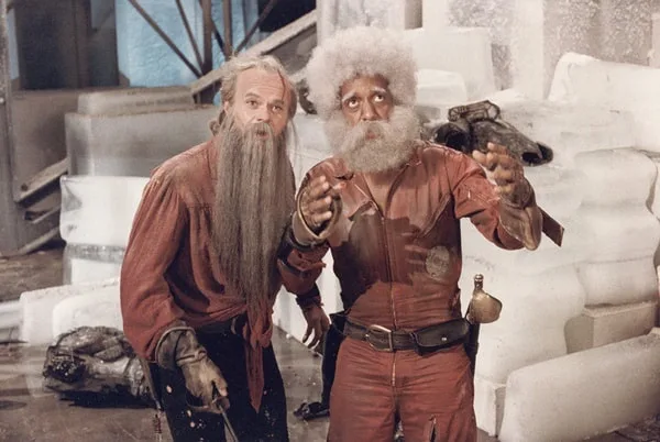 two men in red jumpsuits stand in an ice-filled room, one with a long grey beard and the other with a white afro and beard…