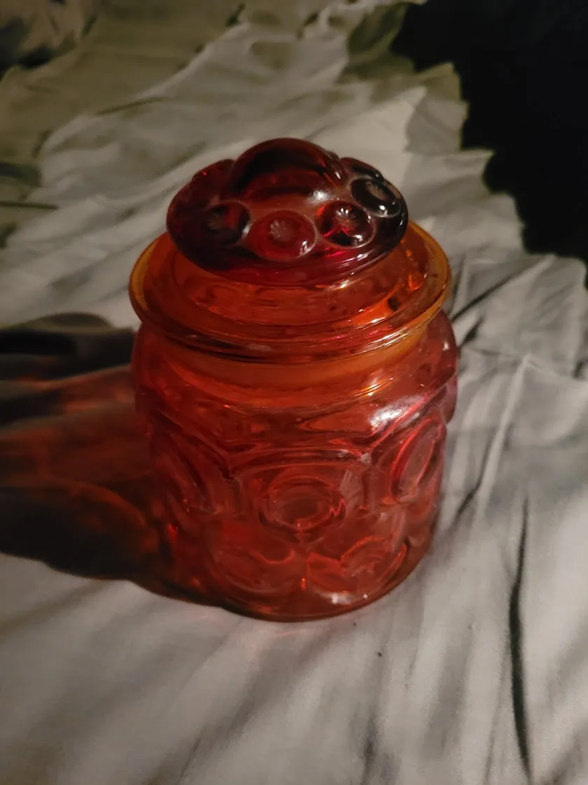 A close-up photo shows a translucent amber-colored glass jar with a decorative lid, sitting on a rumpled white fabric…