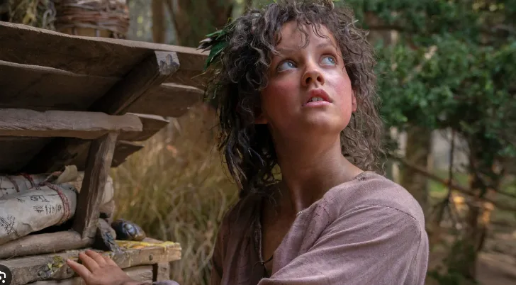 A young girl with curly hair and a leaf crown looks upward with a tearful, pleading expression while standing beside a…