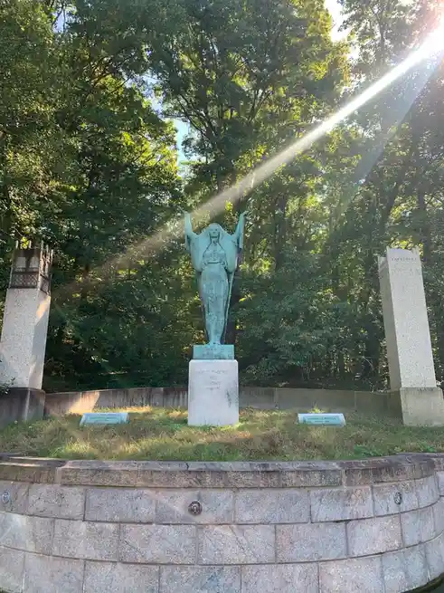 A photograph of a bronze statue of a woman with outstretched arms standing on a stone pedestal in a wooded area, with…