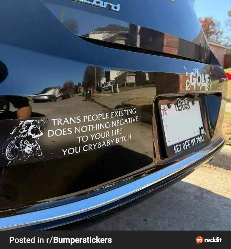 A close-up photo of the rear of a glossy black car showing a bumper sticker with the text 'TRANS PEOPLE EXISTING DOES…
